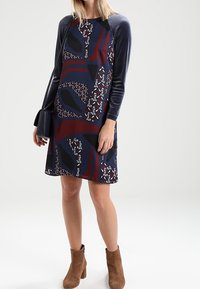 Kleid aus Baumwollmischung mit marineblauem Oberteil und gemustertem Rock in Burgunderrot, Schwarz und Weiß. Lange Ärmel und braune Knöchelstiefel.