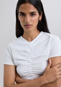 Mujer con cabello largo y oscuro viste una blusa blanca de mangas cortas con fruncidos, cruzando los brazos frente a un fondo gris claro liso.