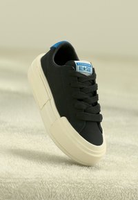 Sneaker nera in tela con suola in gomma bianca, punta arrotondata, lacci neri e un'etichetta sulla linguetta blu e bianca che recita "Converse All-Star."