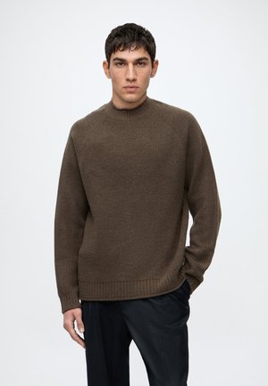 Homme portant un pull en maille marron et un pantalon noir, debout devant un fond clair uni, regardant droit devant.