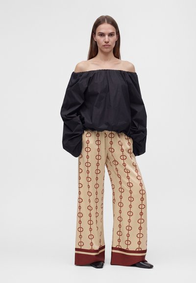 Femme aux longs cheveux raides portant un haut noir ample épaules dénudées et un pantalon large beige avec un motif géométrique rouge.