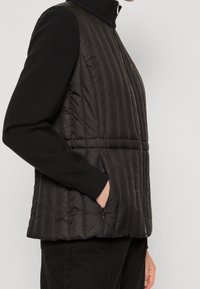 Gilet imbottito nero con colletto alto, texture trapuntata, tasche laterali e dettagli di cucitura verticale. Indossato sopra un maglione nero.