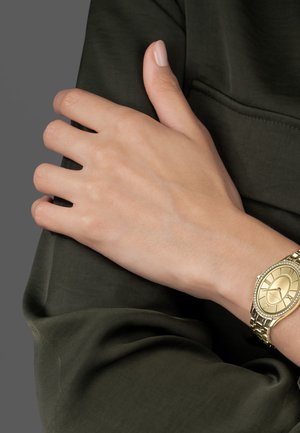 Lijeva ruka odmara na desnoj ruci obučena u tamnozeleni dugih rukava, zlatni ručni sat s ovalnim brojčanikom i metalnom narukvicom.