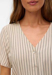 Blusa in lino beige con righe verticali navy, scollatura a V, maniche corte e due bottoni frontali. Tessuto liscio con una vestibilità casual.