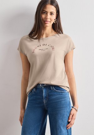 Ung kvinde med langt brunt hår iført beige t-shirt med teksten "Sogni di Capri" og blå jeans, stående mod en ensfarvet væg.