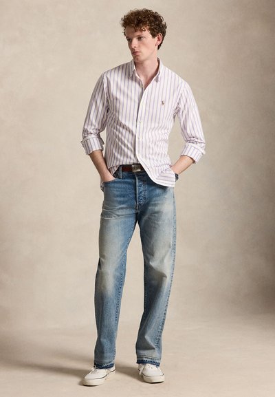 Junger Mann steht mit den Händen in den Taschen, trägt ein hellviolettes gestreiftes Hemd mit Knöpfen, blaue Jeans, weiße Sneakers und eine Armbanduhr.