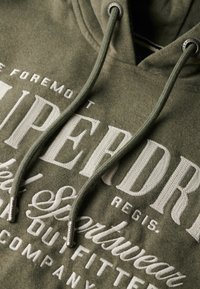 Sudadera verde con un tejido texturizado, que presenta texto bordado en blanco. Incluye cordones con puntas de metal plateado y un corte relajado.