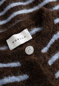 Brązowa, miękka tkanina w jasnoniebieskie paski, biały guzik oraz biała metka odzieżowa z napisem "NOELLA" przyszyta.