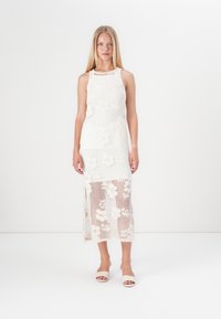 Bela čipkasta obleka s prilegajočim zgornjim delom in polprosojnim prekrivalom, ki ima visoko ovratnik in ravno silhueto. Bež natikači.