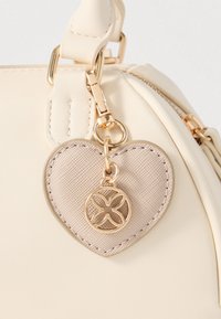 Crème faux-lederen tas met gouden hardware. Heeft een afneembare hartvormige hanger met een getextureerd oppervlak en een circulair logodetail.