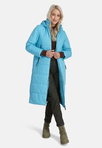 Gai zils pūkains mētelis ar kapuci, ar pārsistām šuvēm, ar rāvējslēdzēju un sānu kabatām, uzvilkts virs brūna tops un tumši zaļiem biksēm.