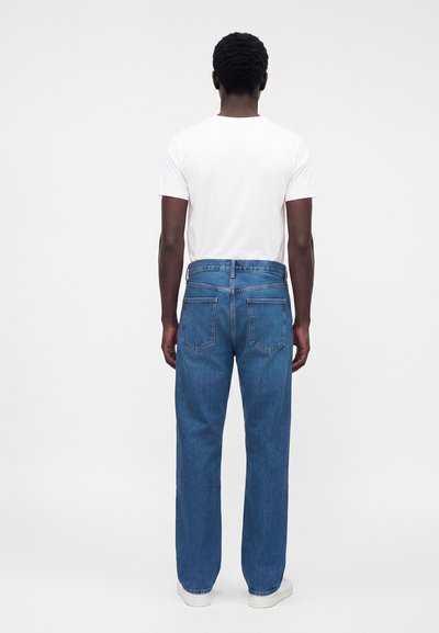 Homme de dos portant un t-shirt blanc, un jean droit bleu et des baskets blanches sur un fond clair uni.