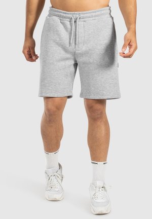 Man in lichte grijze drawstring shorts, met witte crew- sokken en witte sneakers, staande tegen een effen lichte achtergrond.