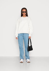 Sweatshirt branco com logótipo texturizado, jeans de perna reta azul claro, mala preta, ténis brancos com pormenores castanhos e óculos de sol escuros.