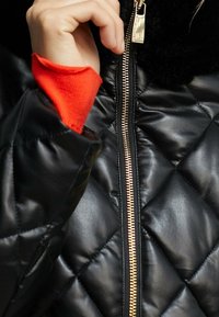 Svart vadderad pufferjacka med gulddragkedja, faux fur-krage och röda stickade manschetter som syns under jackans ärm.