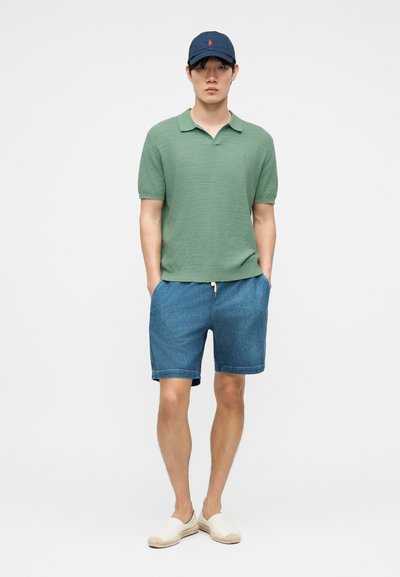 Νέος άνδρας που φοράει πράσινο κοντομάνικο πόλο, μπλε τζιν σορτς, σκούφο ναυτικού χρώματος και λευκά παπούτσια slip-on, στέκεται με τα χέρια στις τσέπες.