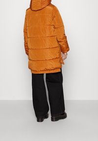 Oranssi puffertakki hupulla, jossa on vaakasaumat, rento malli ja hieman kiiltävä pinta, yhdistettynä mustiin leveälahkeisiin housuihin.