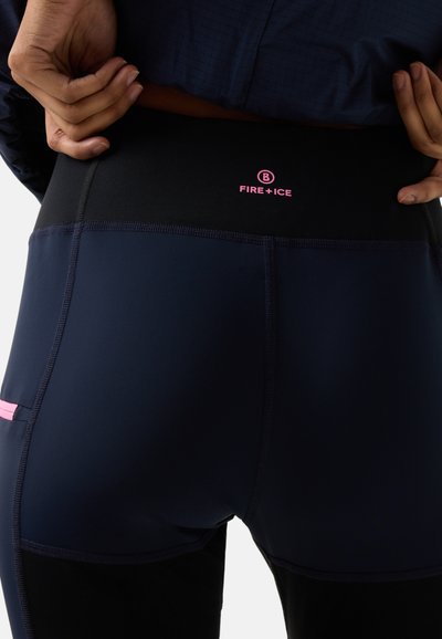 Hænder, der justerer linningen på mørkeblå atletikleggings med "FIRE+ICE"-logo i pink på bagsiden.