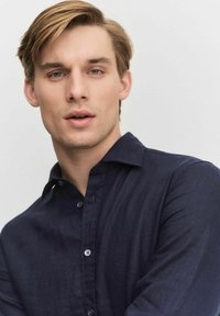 Marineblauwe button-up shirt met een zachte textuur, kraag en zichtbare knopen; gedragen door een model met licht haar en een neutrale achtergrond.