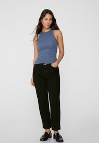 Blau gerippter Tanktop mit hohem Ausschnitt, kombiniert mit hochgeschnittenen schwarzen Hosen und schwarzen Slippern. Das Model steht mit einer Hand in der Tasche.