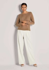 Frau in einem braunen Strickpullover, weißen weiten Hosen, beigen High Heels und dekorativen Ohrringen, die vor einem einfachen hellen Hintergrund steht.