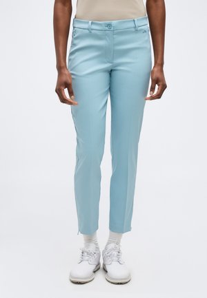 Άτομο που φοράει στενά παντελόνια ανοιχτού μπλε χρώματος, μπεζ μπλούζα, λευκές κάλτσες και λευκά αθλητικά παπούτσια, στέκεται μπροστά σε απλό φόντο.