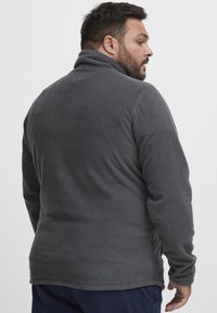 Dunkelgraue Fleecejacke mit einem hohen Kragen, strukturiertem Stoff und langen Ärmeln. Bietet einen lockeren Sitz und seitliche Belüftungen für zusätzlichen Komfort.