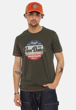 Jeune homme portant un t-shirt olive Von Dutch et une casquette orange Von Dutch, avec des tatouages sur les bras, posant devant un fond uni.