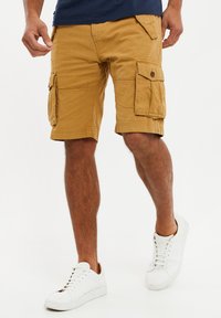 Cargo-Shorts in einem senfgelben Farbton aus einem weichen, strapazierfähigen Stoff. Mit Seitentaschen, Knopfdetails und einer lockeren Passform. Kombiniert mit weißen Turnschuhen.