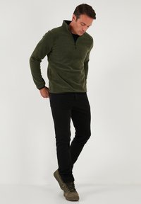 Olivengrøn fleece-pullover med kvart-lynlås-krave. Matchet med tætsiddende sorte jeans og grønne vandrestøvler, hvilket viser et afslappet look.