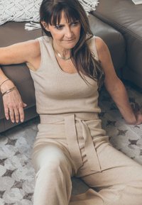 Beige, geripptes Tanktop, kombiniert mit hochgeschnittenen, mit Schleife versehenen Hosen aus weichem Material, sitzend auf einem gemusterten Teppich vor einer neutralen Ledercouch.