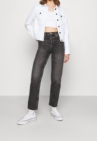 Mujer vistiendo una chaqueta corta blanca, un top corto blanco, jeans negros de cintura alta y zapatillas altas blancas, de pie frente a un fondo liso.