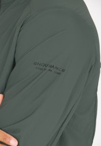Grøn letvægtsjakke med en struktureret overflade, udstyret med et sort logo med teksten "ENDURANCE HERO OF THE ROAD" på ærmet.