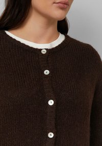 Brauner gestrickter Cardigan mit strukturierter Oberfläche, der vorne sechs runde, weiße Knöpfe und einen gerippten Halsausschnitt hat.