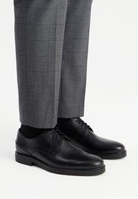 Sorte læderdressko med en glat overflade, med en rund tå og fem snørrehuller, parret med grå ternede bukser.