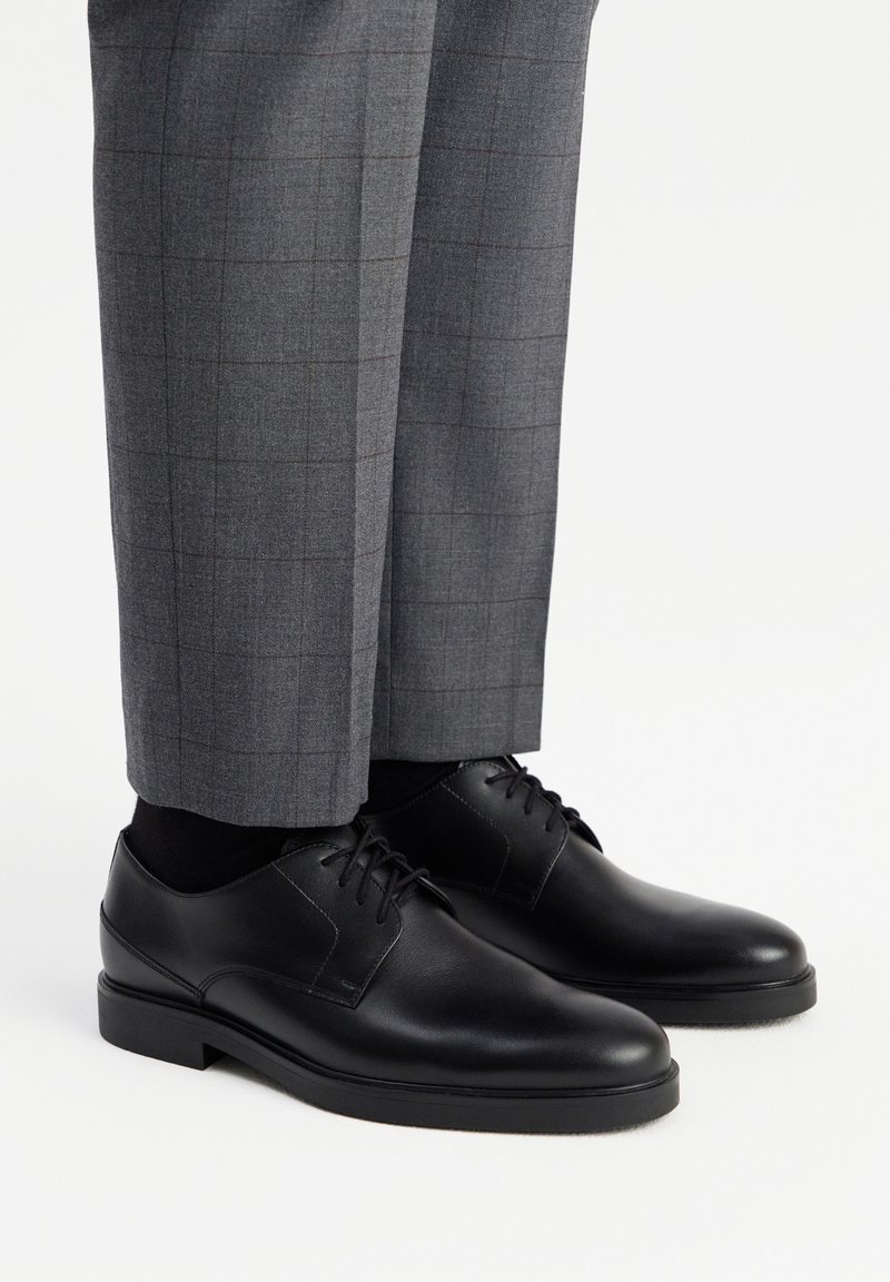 Sorte læderdressko med en glat overflade, med en rund tå og fem snørrehuller, parret med grå ternede bukser.