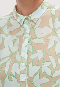 Primo piano di una camicia con colletto su sfondo beige e motivi astratti di foglie azzurre contornati di verde.