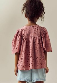 Rosa Augenlederbluse mit Puffärmeln, die floral geschnittene Aussparungen und eine Knopfleiste auf der Rückseite aufweist, kombiniert mit hellblauen Streifenshorts.