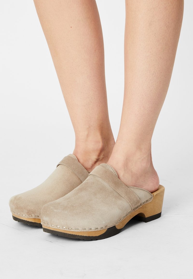 Pieds portant des sabots en daim beige à enfiler avec semelles en bois et talons bas, présentés sur fond blanc.