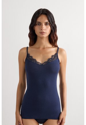 Frau mit welligem dunklem Haar, die ein marineblaues Camisole mit Spitzenbesatz und passende Unterwäsche vor einem schlichten weißen Hintergrund trägt.