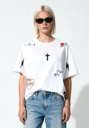 Femme aux cheveux blonds courts portant des lunettes de soleil noires, un t-shirt blanc avec des imprimés graphiques noirs et rouges, et un jean bleu clair.
