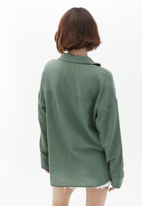 Camicia verde oversized con bottoni, realizzata in un tessuto testurizzato, dotata di colletto e maniche lunghe. La vista dal retro mostra un orlo curvo.