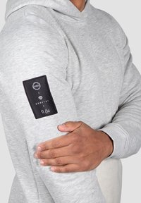 Sudadera gris con una textura suave, que presenta un parche negro en la manga con un logo y detalle de firma. Los puños acanalados completan el diseño.