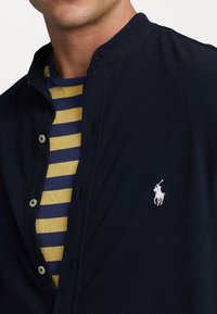 Granatowa koszula z guzikami i stójką, z białym logo, nałożona na żółtą i granatową koszulę w paski. Teksturowany materiał.