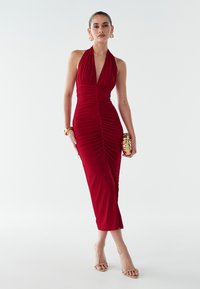 Moteris stovi šviesiai raudono, raukinio formos, be rankovių, halter stiliaus suknelėje, laiko auksinį rankinuką, dėvi auksines auskarų žiedus ir atitinkantį apyrankę.