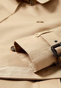 Beige trenčkot vyrobený z texturované látky má složený rukáv s knoflíkem, nastavitelné poutko a kovové doplňky.