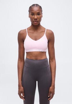 Femme en forme avec des cheveux tressés, portant un soutien-gorge de sport rose clair et un legging gris foncé taille haute, debout devant un fond uni.