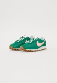 Žalios tinklinės ir zomšinės sportbačiai su šviesiai rožiniu swoosh, baltais raišteliais ir rudais guminiais padu. Turintys klasikinį bėgimo batelių dizainą.