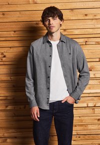 Camicia grigia con bottoni, dal taglio rilassato sopra una t-shirt bianca, abbinata a jeans in denim scuro. Presenta una trama sottile e un piccolo dettaglio con logo.