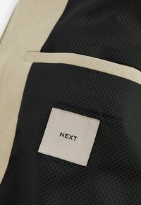 Tissu noir texturé avec une étiquette beige cousue affichant le mot « NEXT » en lettres majuscules noires. Coutures et bordure beiges visibles.